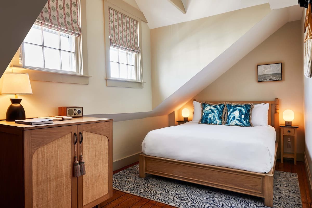 A bedroom with a wooden bed, a desk with a lamp, a radio, and a window with patterned curtains.