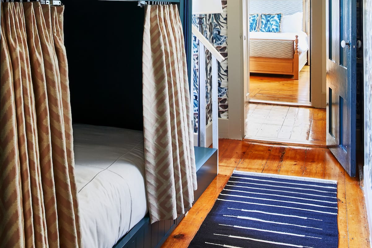 A room with bunk beds, a patterned wallpaper, a wooden floor, and a view into another room with a bed and a blue door.