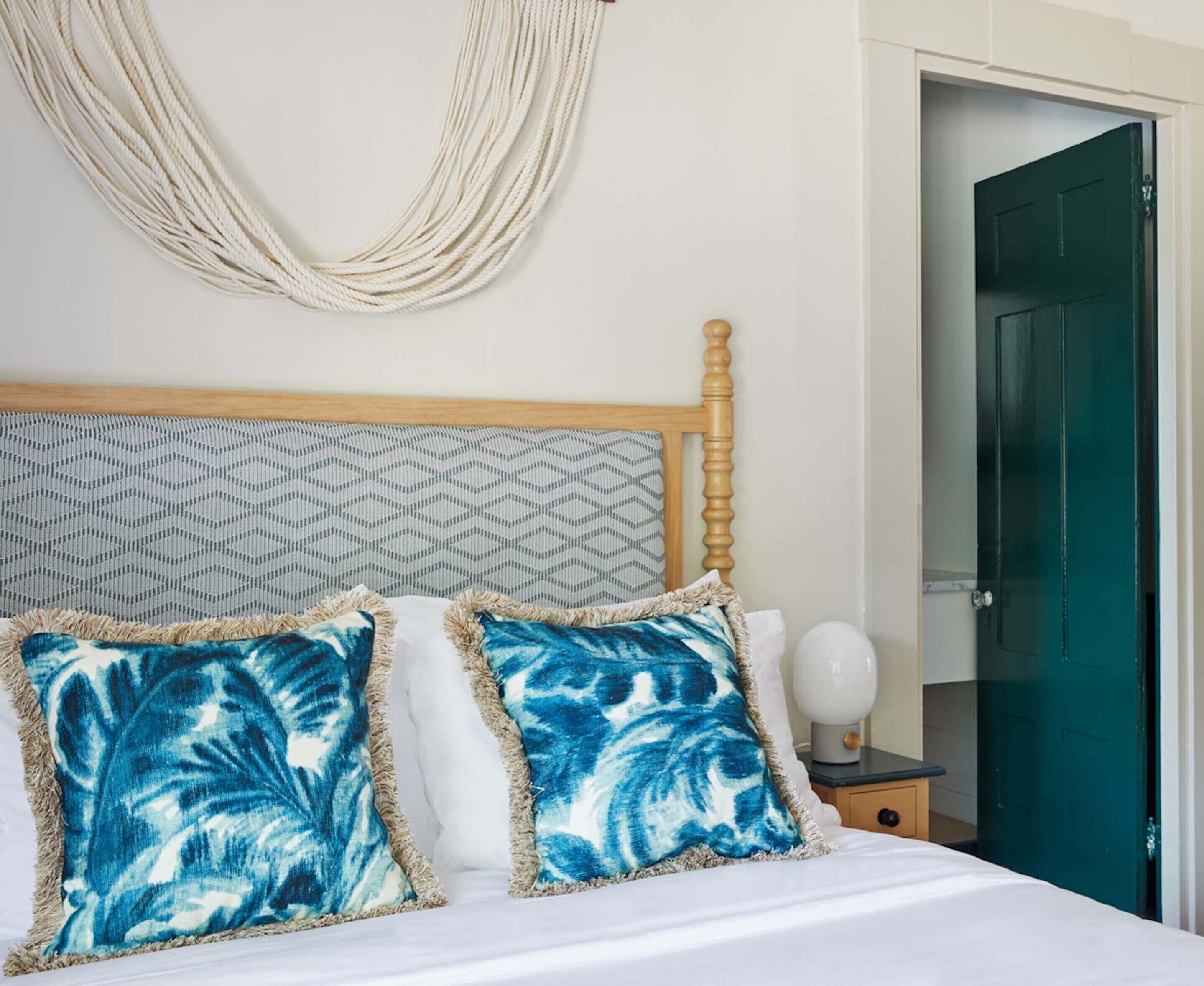 Una cama con dos almohadas azules estampadas, un cabecero de madera, un tapiz decorativo en la pared y una puerta verde al fondo.
