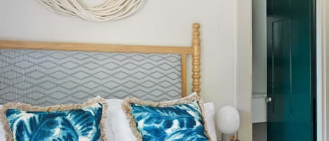 Una cama con dos almohadas azules estampadas, un cabecero de madera, un tapiz decorativo en la pared y una puerta verde al fondo.