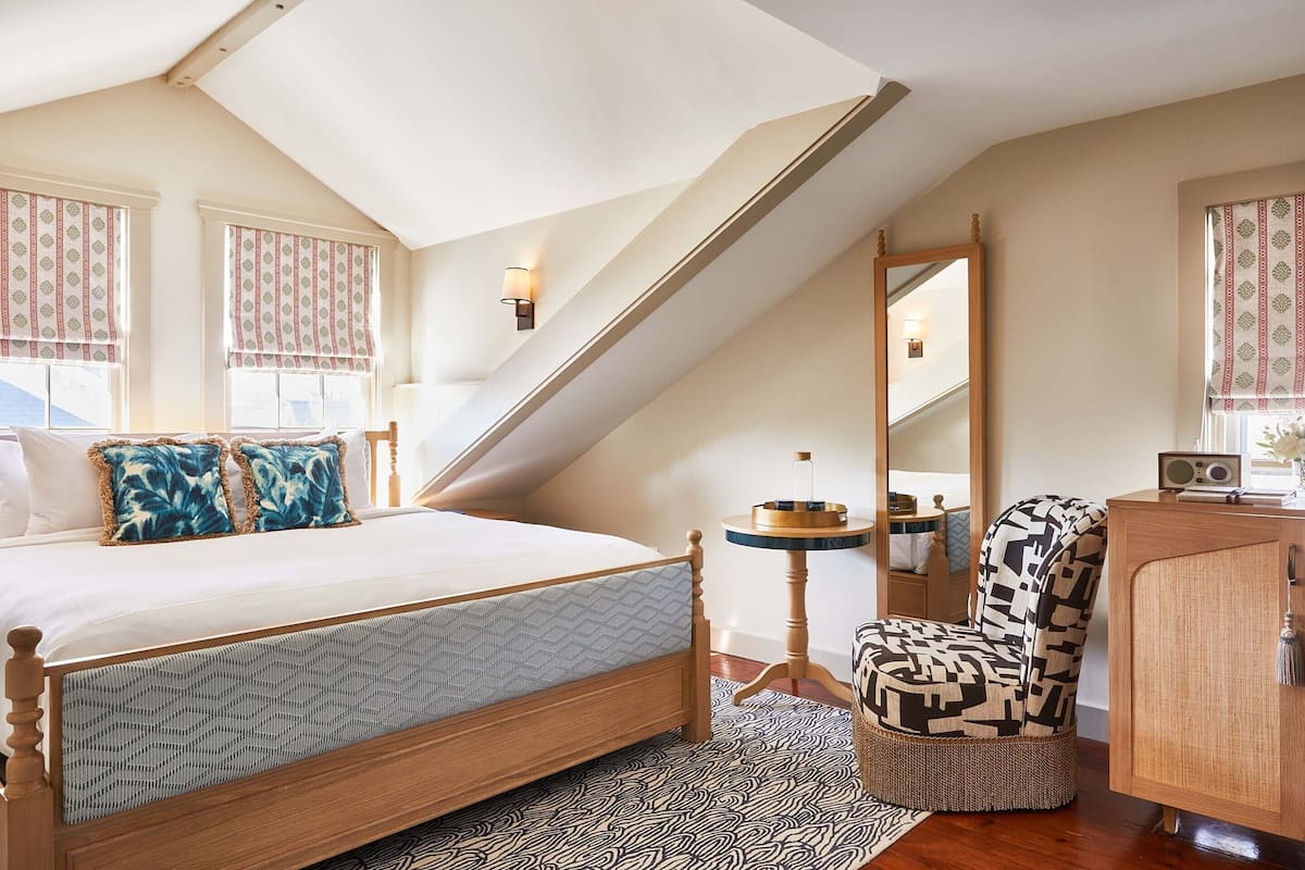 A bedroom with a sloped ceiling, a wooden bed, patterned pillows, a round side table, a chair, a mirror, and a cabinet.