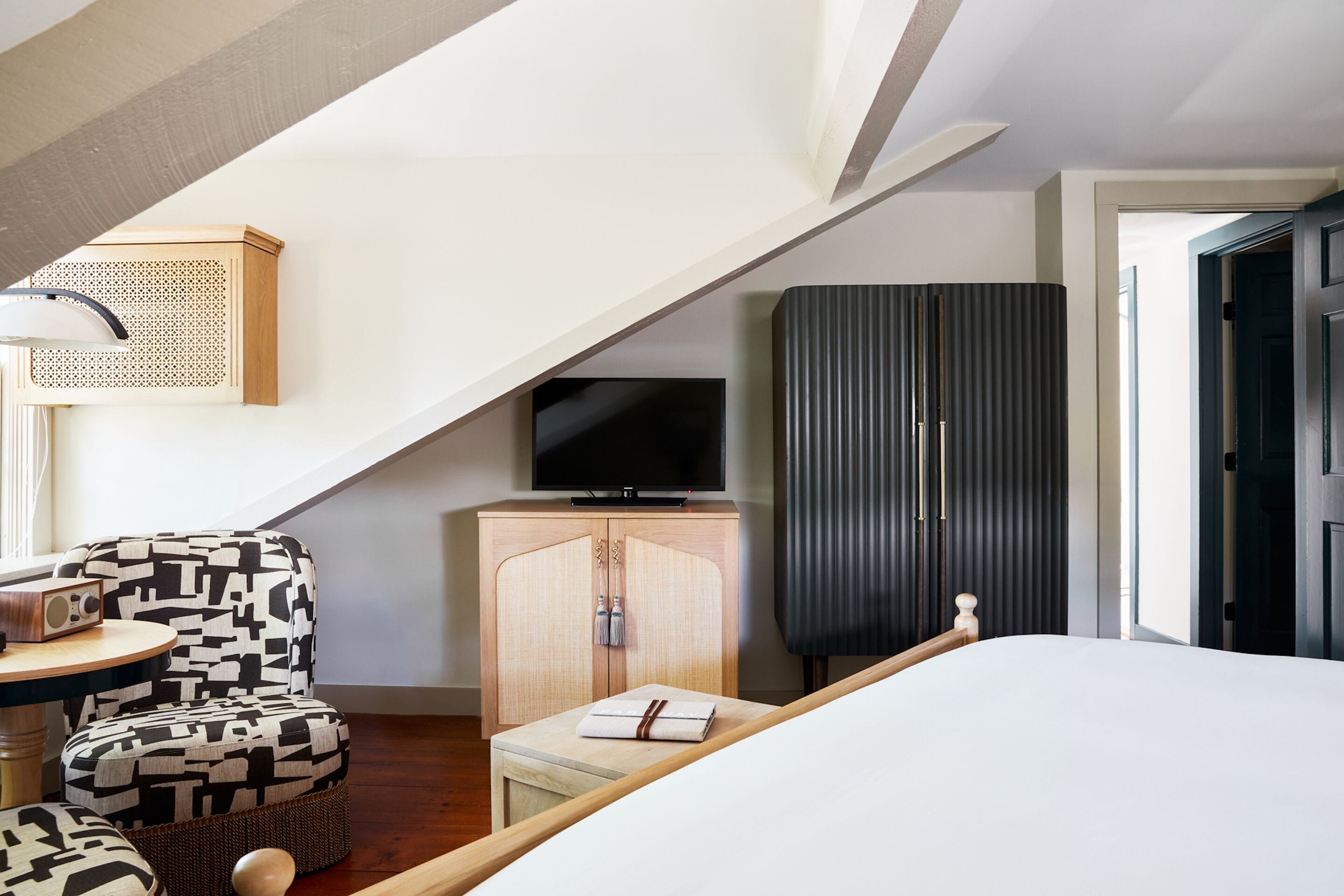 A modern hotel room with a bed, a television, a wooden cabinet, a patterned armchair, and a small table with a radio.