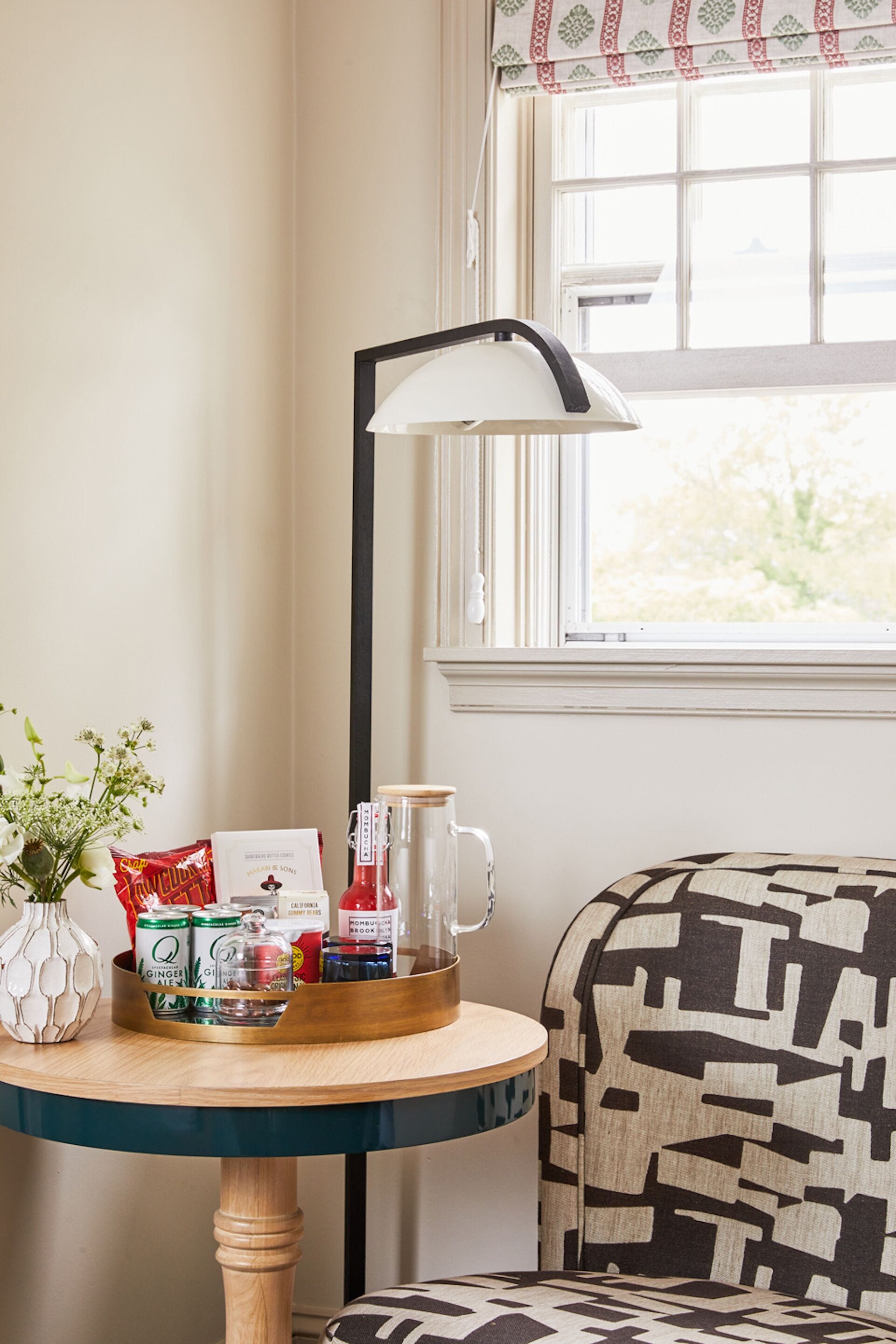 A table with a tray of snacks, a mug, and a bottle, a patterned sofa, and a lamp near a window.