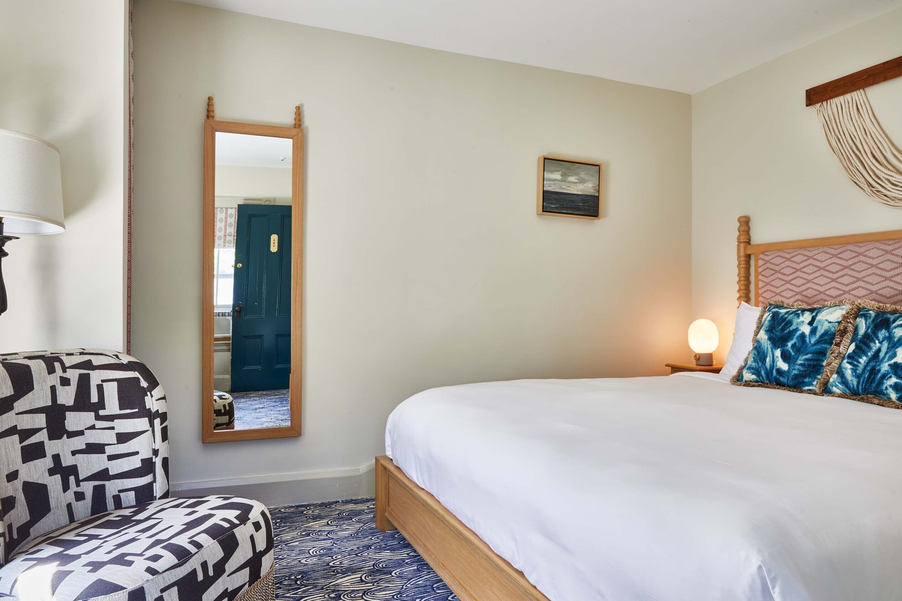 A neatly arranged bedroom with a large bed, a chair, a mirror, and a small picture on the wall.