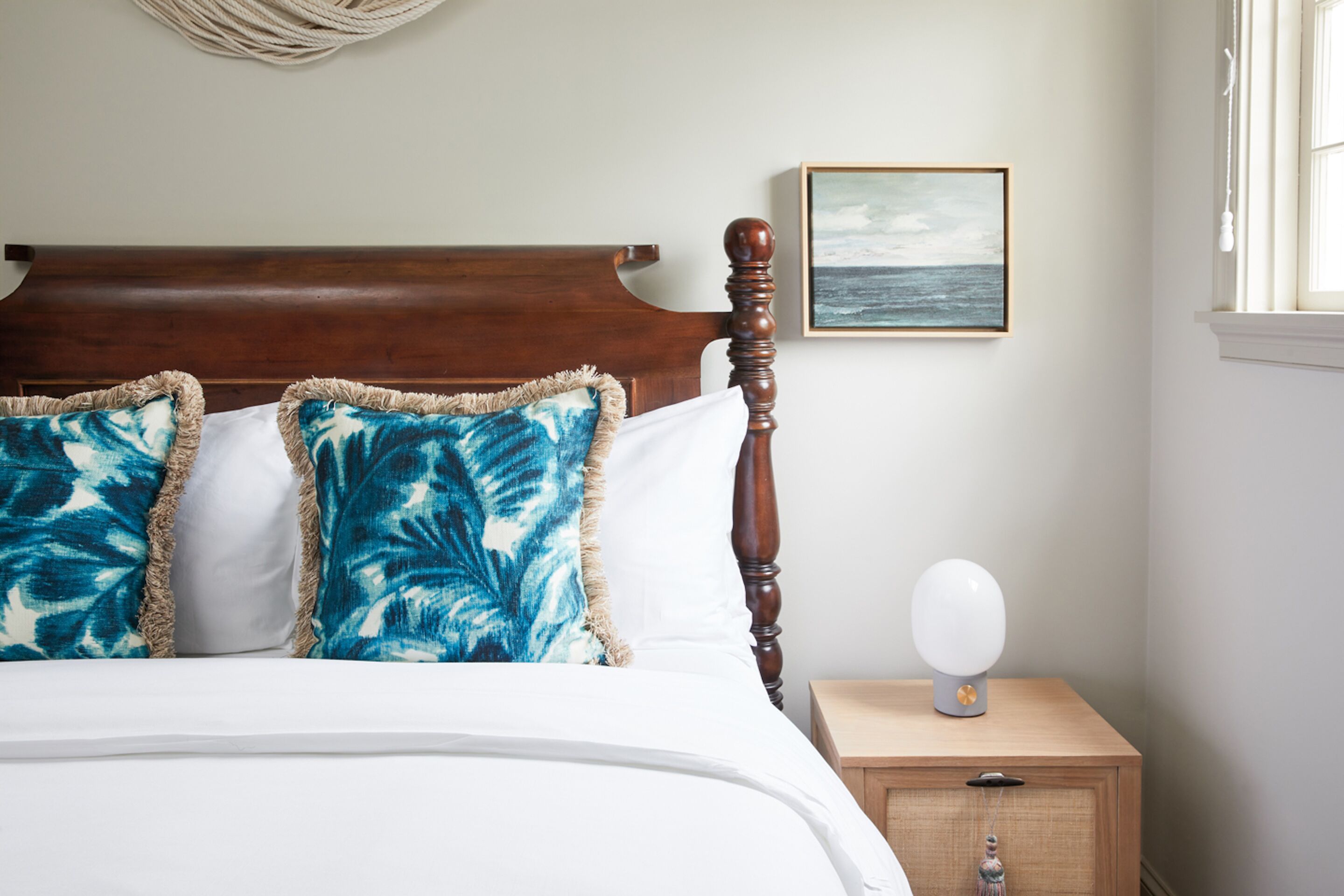 A bed with a wooden headboard, two pillows with a blue leaf pattern, a bedside table with a white lamp, and a framed picture of the ocean on the wall.