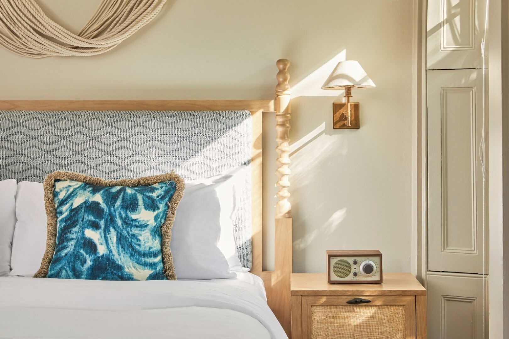 Una habitación de hotel con una cama, una mesita de noche con una radio, una lámpara de pared y un adorno colgante.