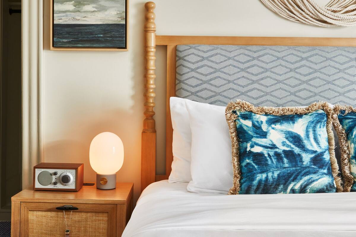 A neatly made bed with a patterned pillow, a bedside table with a lamp, and a wall-mounted macrame piece.