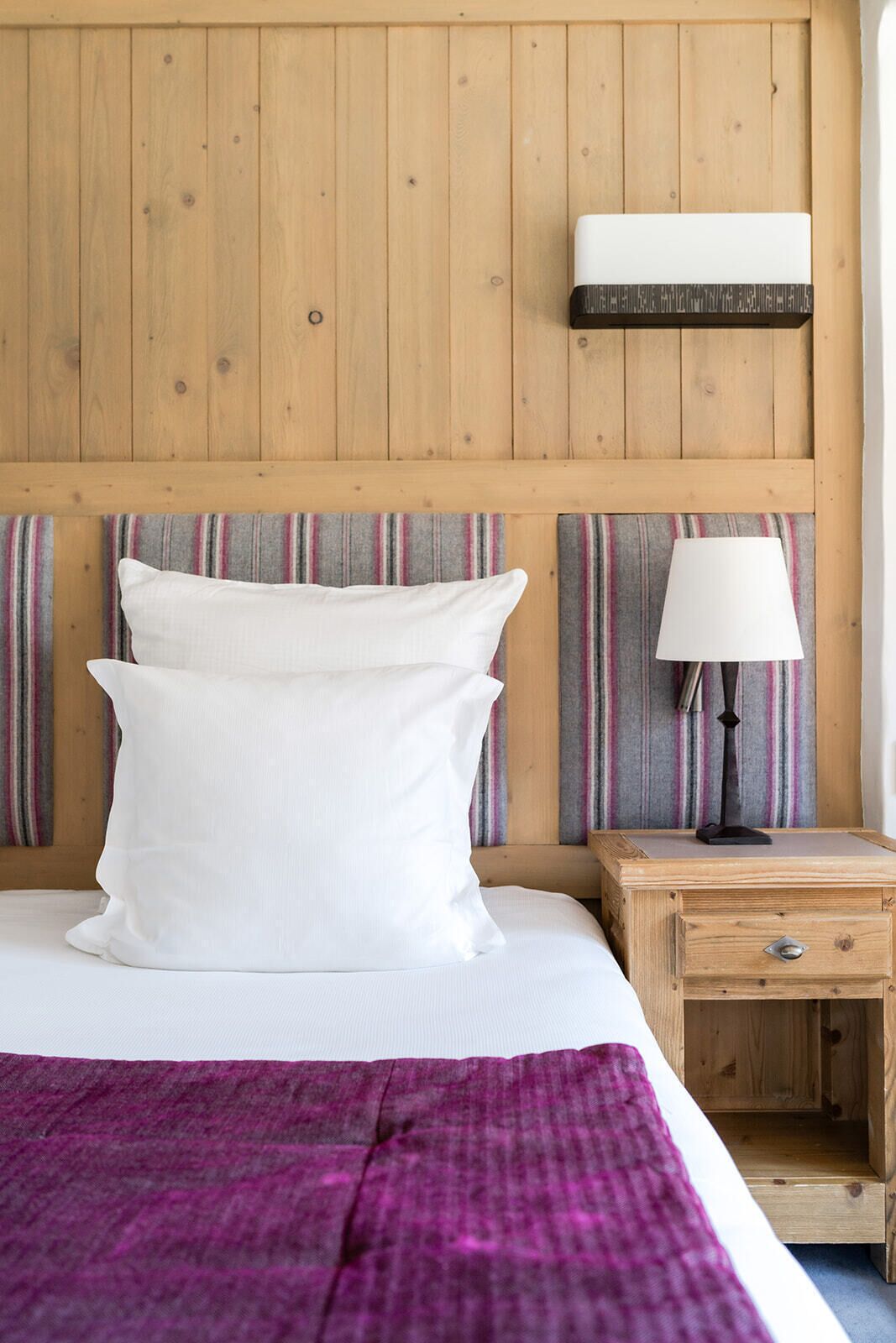 A bed with white bedding, a purple blanket, and two pillows. There is a wooden nightstand with a lamp and a small drawer. The wall is wooden with a horizontal panel above the bed.