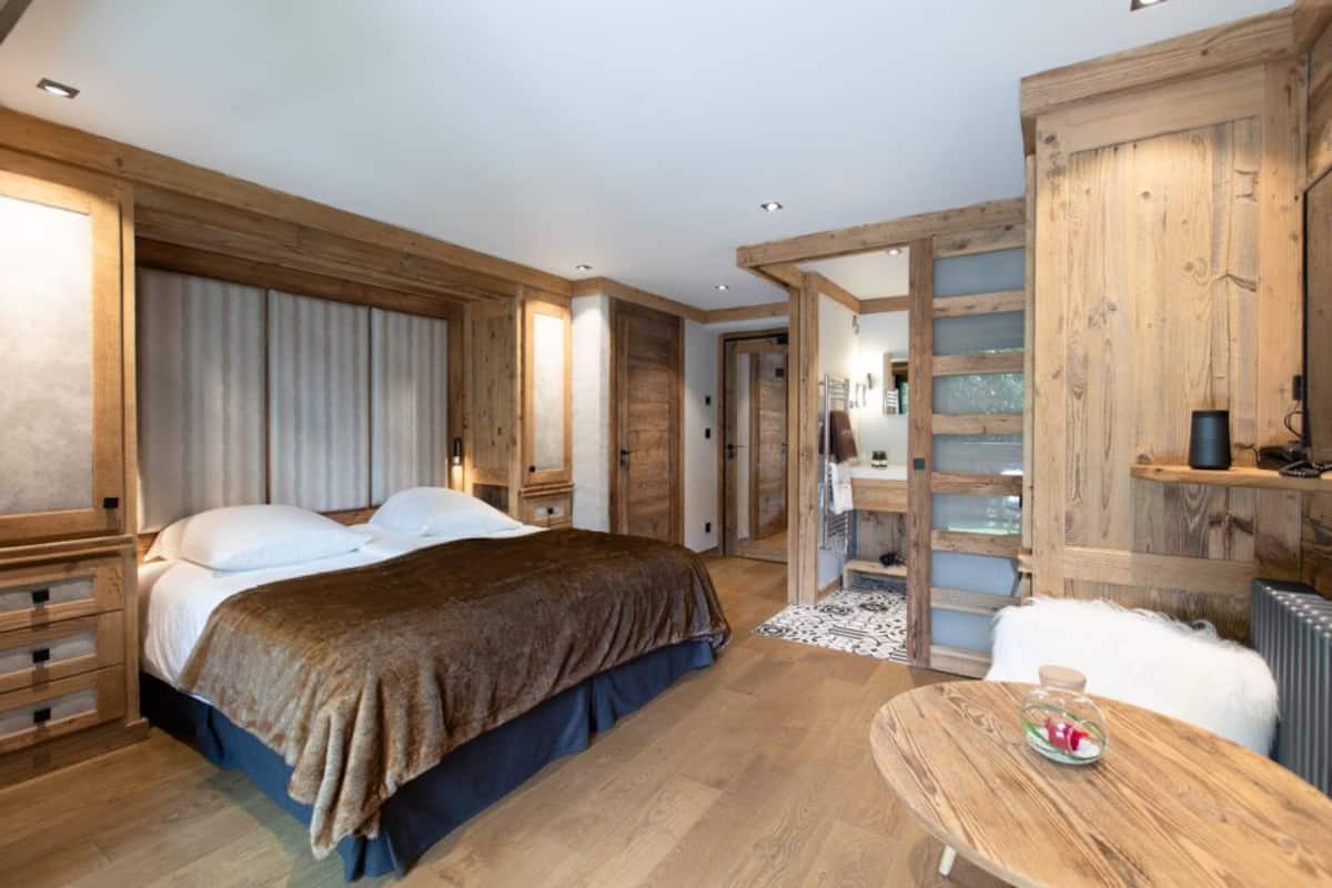 A bedroom with a large bed, wooden walls, a small table, and a bathroom visible through a glass door.