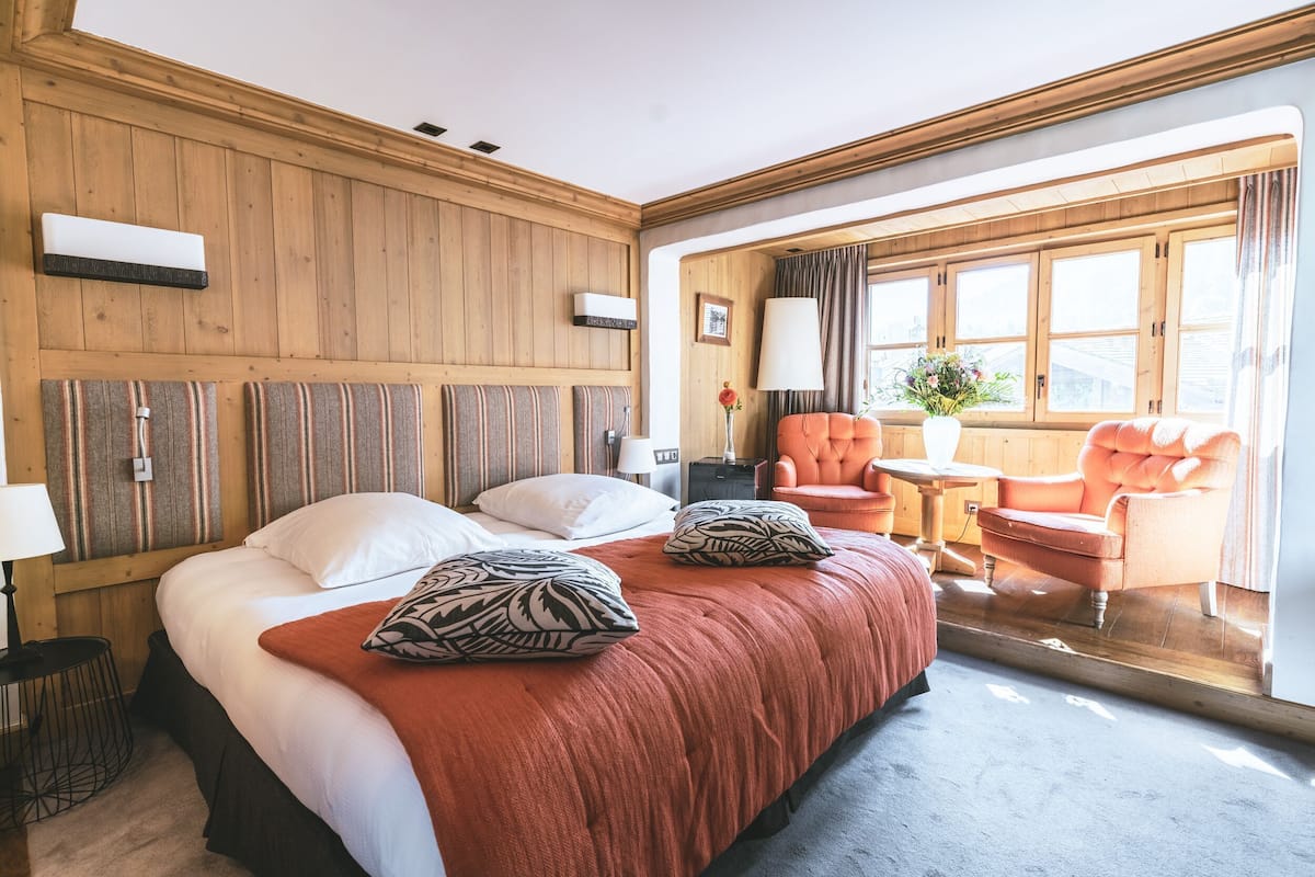 A hotel room with a wooden paneled wall, a large bed with a red blanket, two pillows, a nightstand, a lamp, a chair, and a window with curtains.