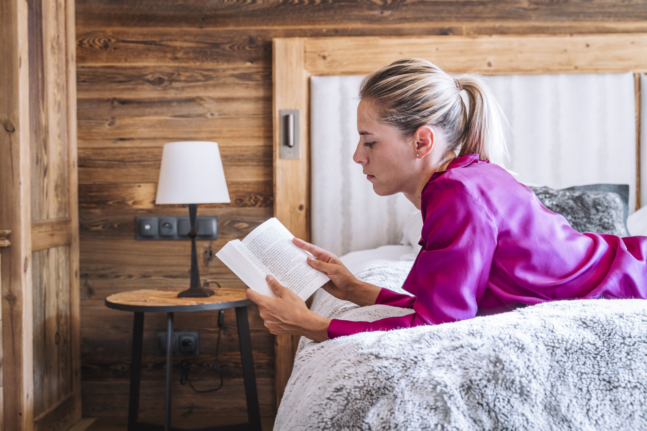 A person reading a book in a room with wooden walls, a small table with a lamp, and a bed with a white blanket.
