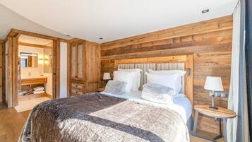 A bedroom with a wooden headboard, a large bed with multiple pillows, a bedside table with a lamp, and a bathroom visible through an open door.
