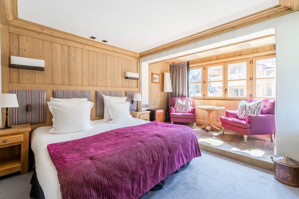A hotel room with a large bed, a wooden headboard, a small table, a pink armchair, and a view of the outside through large windows.