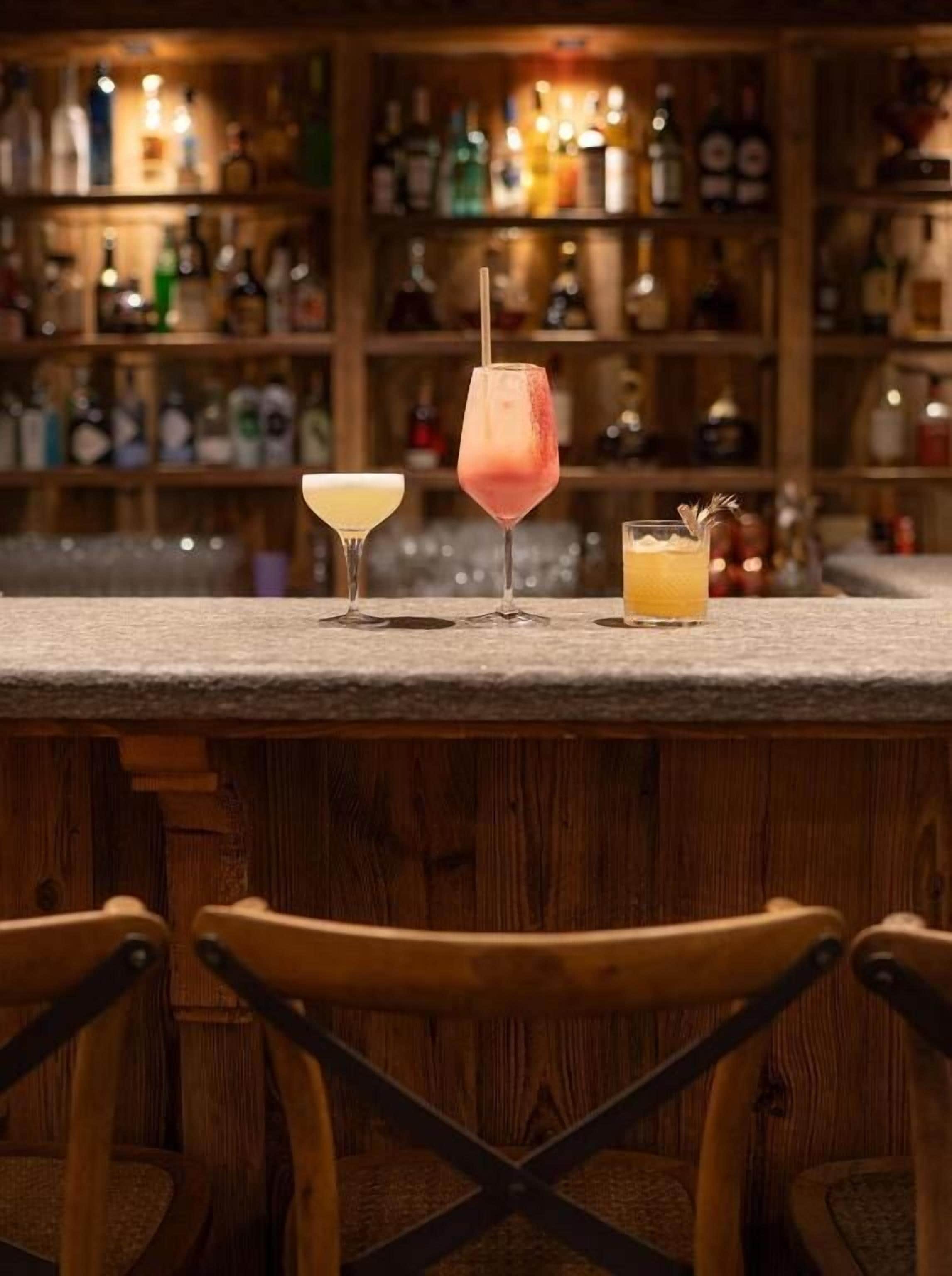 A bar area with three drinks on the counter and wooden chairs in the foreground.