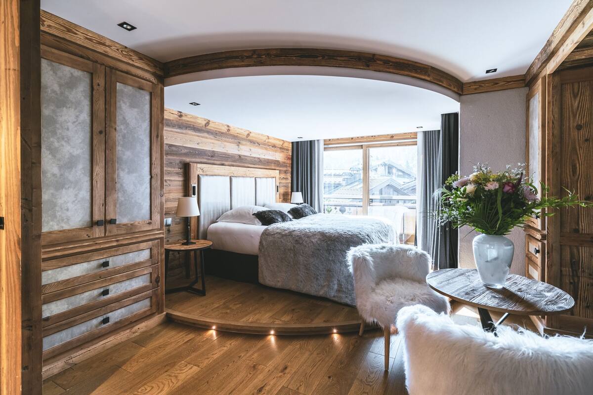 A cozy bedroom with a wooden ceiling, a bed with a fluffy blanket, a small round table with a vase of flowers, and a fluffy chair.