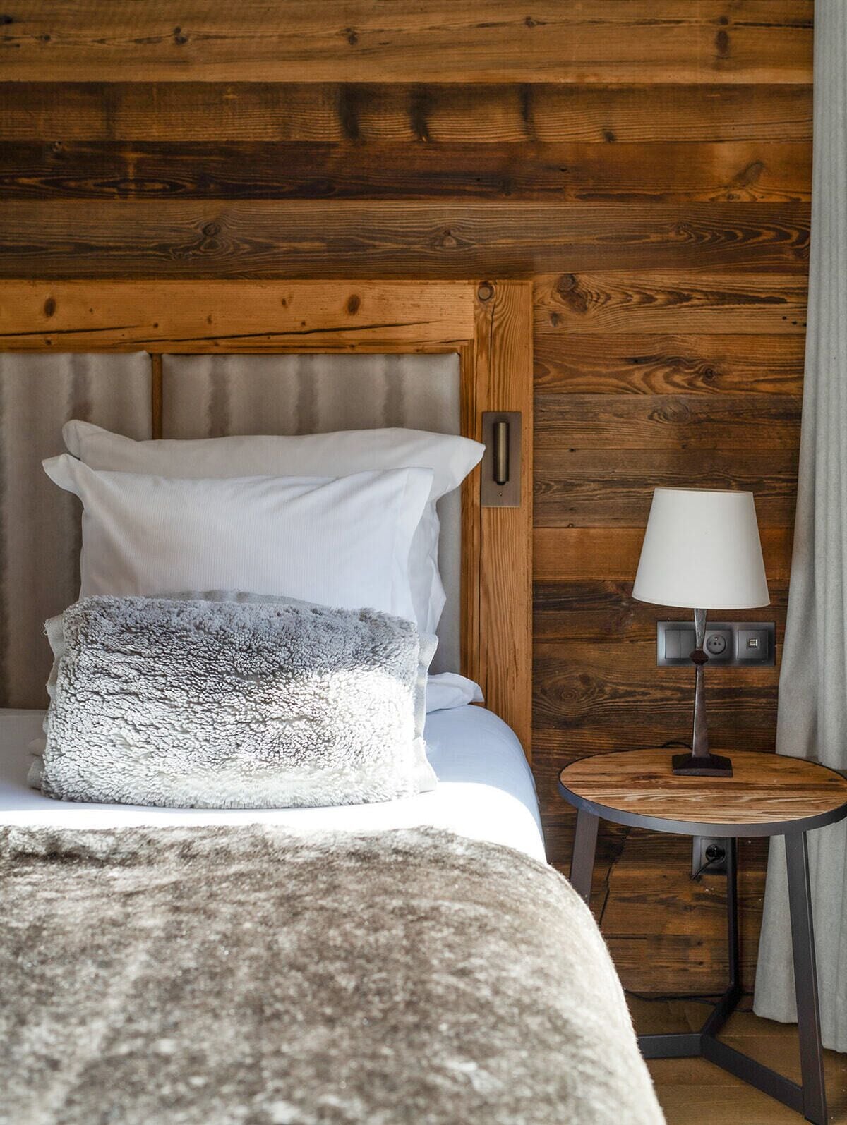 A neatly made bed with white and grey pillows, a round table with a lamp, and wooden paneling on the walls.