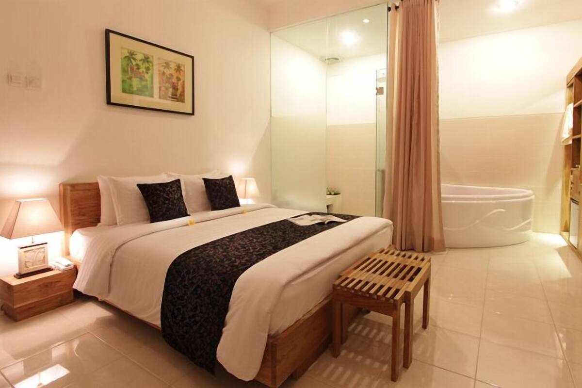 A hotel room with a bed, bedside table, lamp, and a shower area with a bathtub.