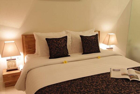 A neatly made bed with a white duvet and two patterned pillows, flanked by bedside tables with lamps and a framed picture. - The Adnyana Villas & Spa (Tibubeneng)
