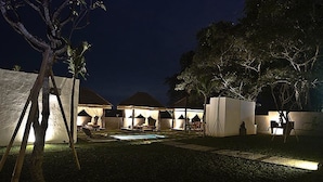Vista nocturna de un resort con varias cabañas sobre el agua, una piscina y un área exterior bien iluminada.