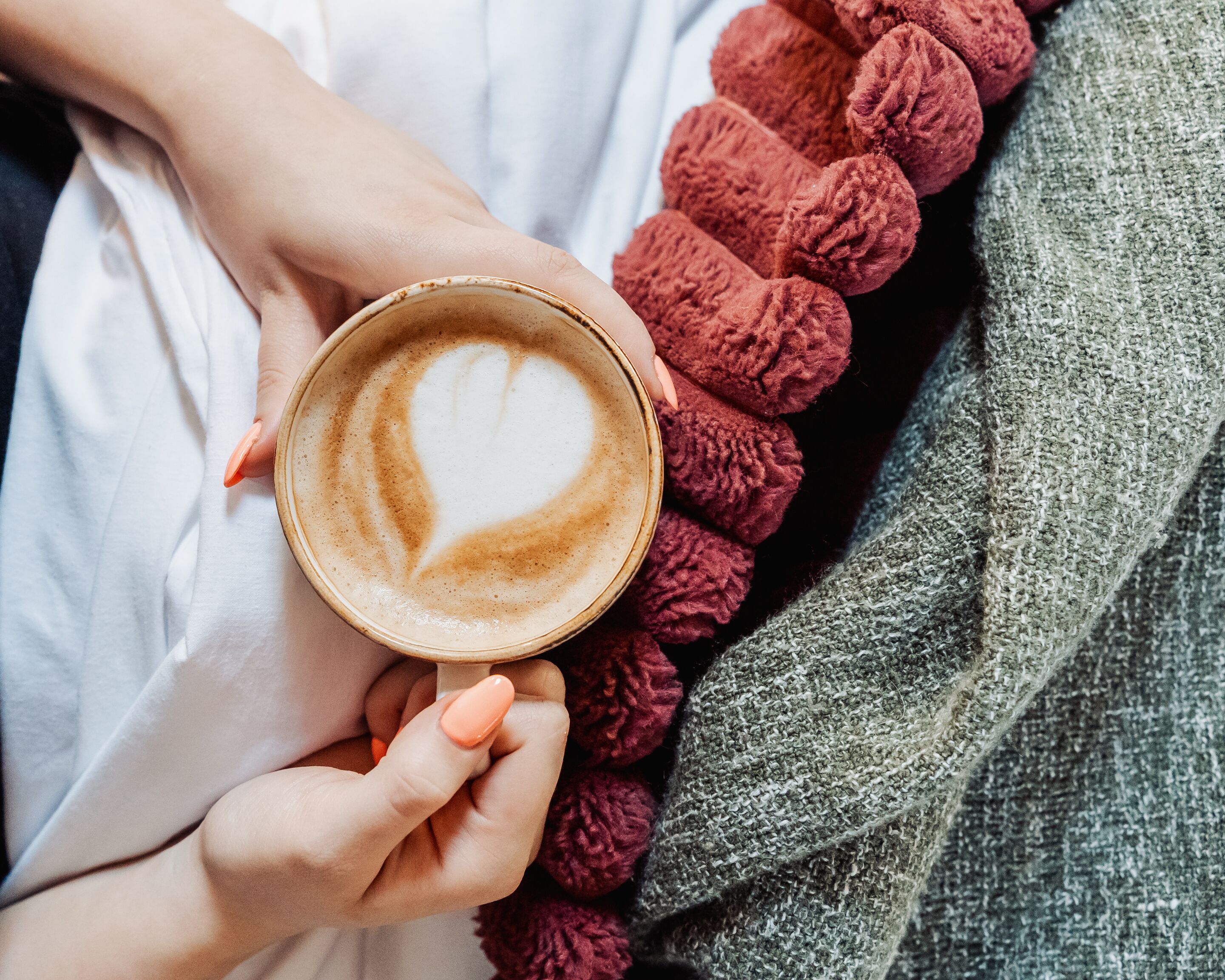 Iemand houdt een koffiekop vast met hartvormig latte-art, draagt een witte kledingstuk en een gebreid rood accessoire.