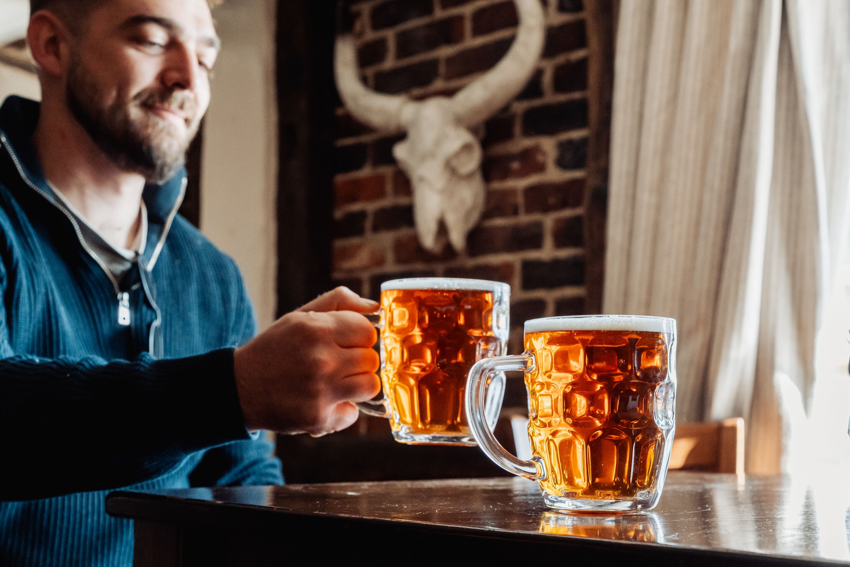 Eine Person hält ein Bierglas, ein weiteres Glas steht auf dem Tisch und eine Backsteinmauer mit einer Kuhskulls Dekoration.