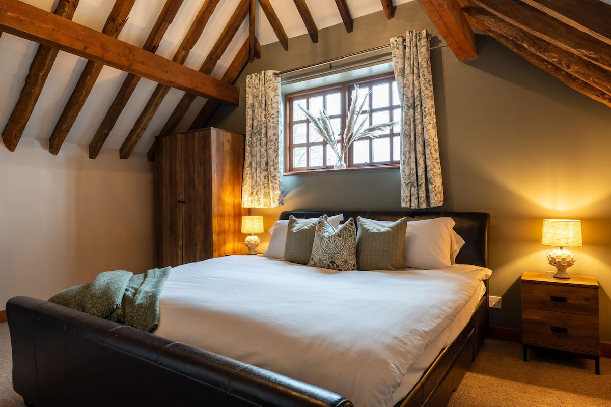 A bedroom with a wooden ceiling, a large bed with white linens, patterned pillows, a nightstand with a lamp, and a window with curtains.