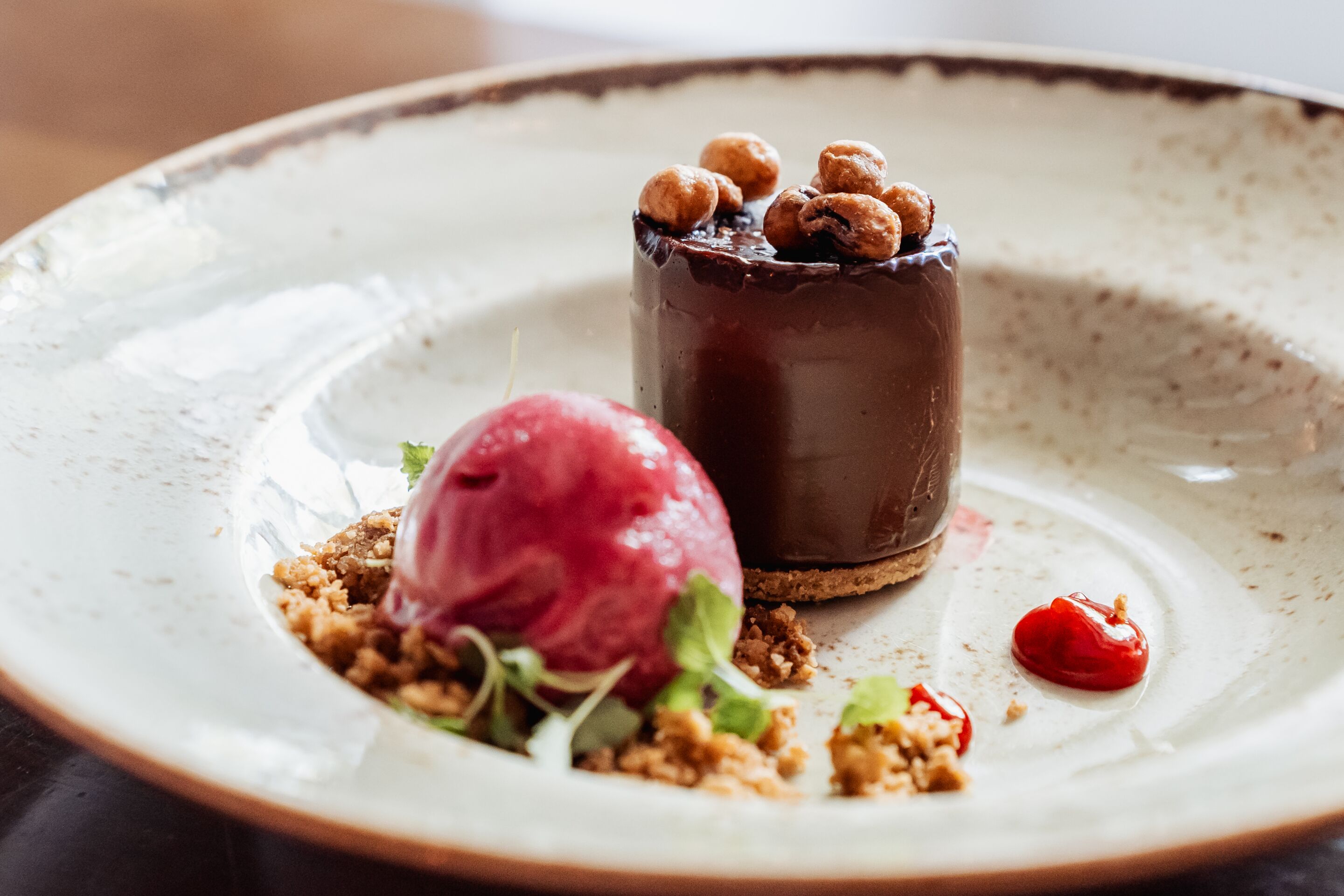 A dessert with a chocolate dome, nuts, and a scoop of ice cream on a plate.