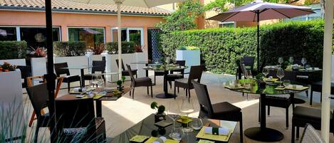 Un área de comedor al aire libre con mesas y sillas, sombrillas y vistas a un edificio y vegetación.
