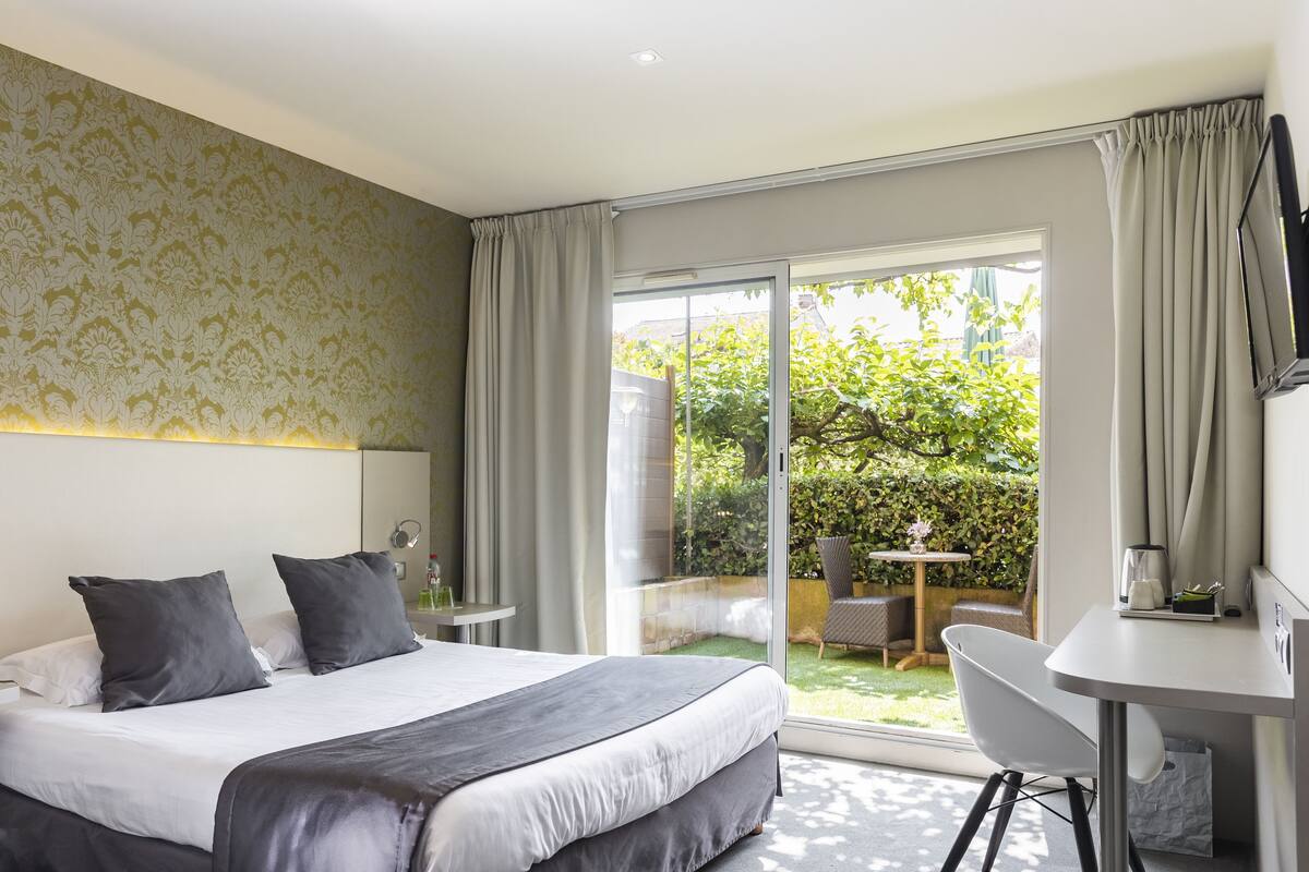 A modern hotel room with a large bed, a desk, and a view of a garden through a sliding glass door.