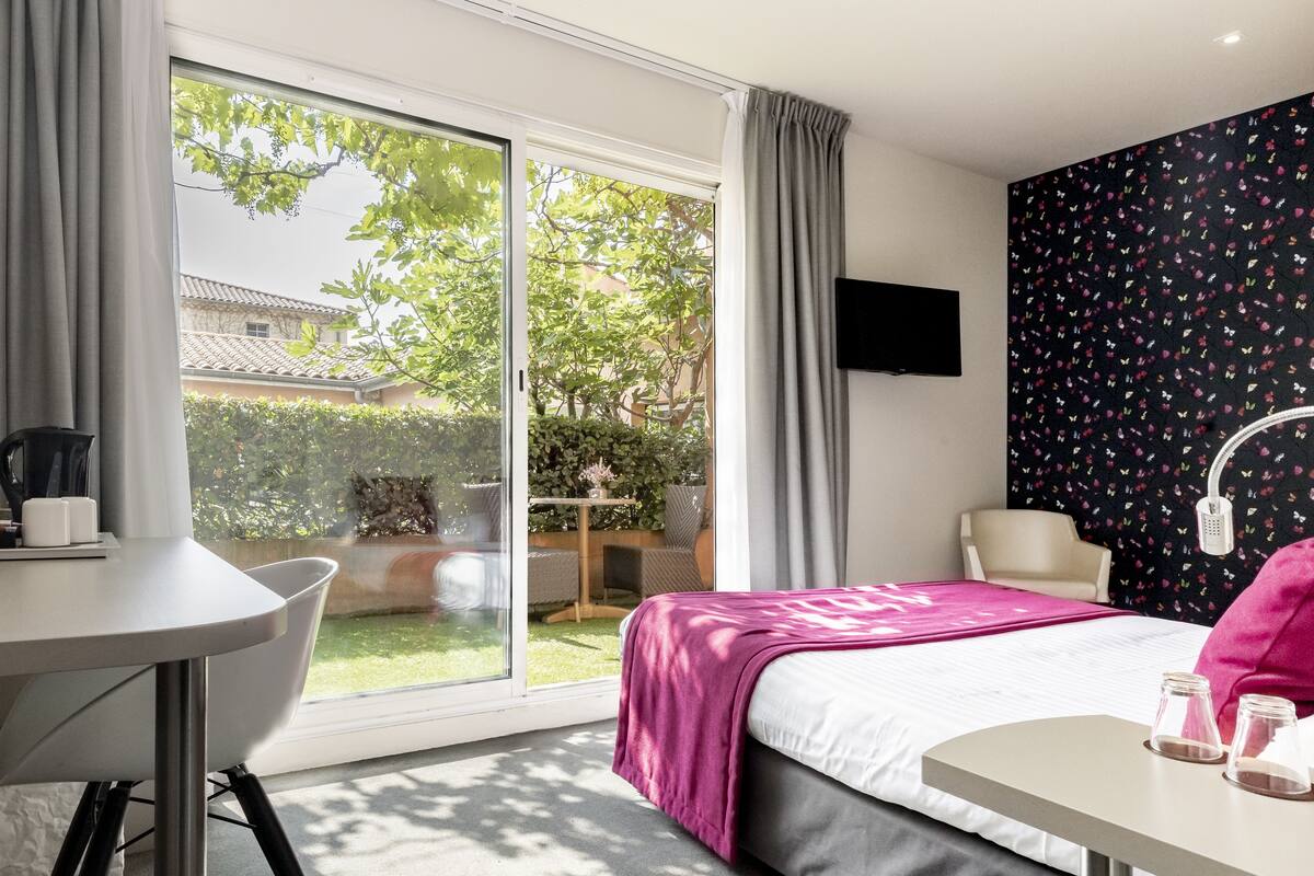 A hotel room with a bed, a table, a chair, a TV, and a balcony with a view of greenery and buildings.