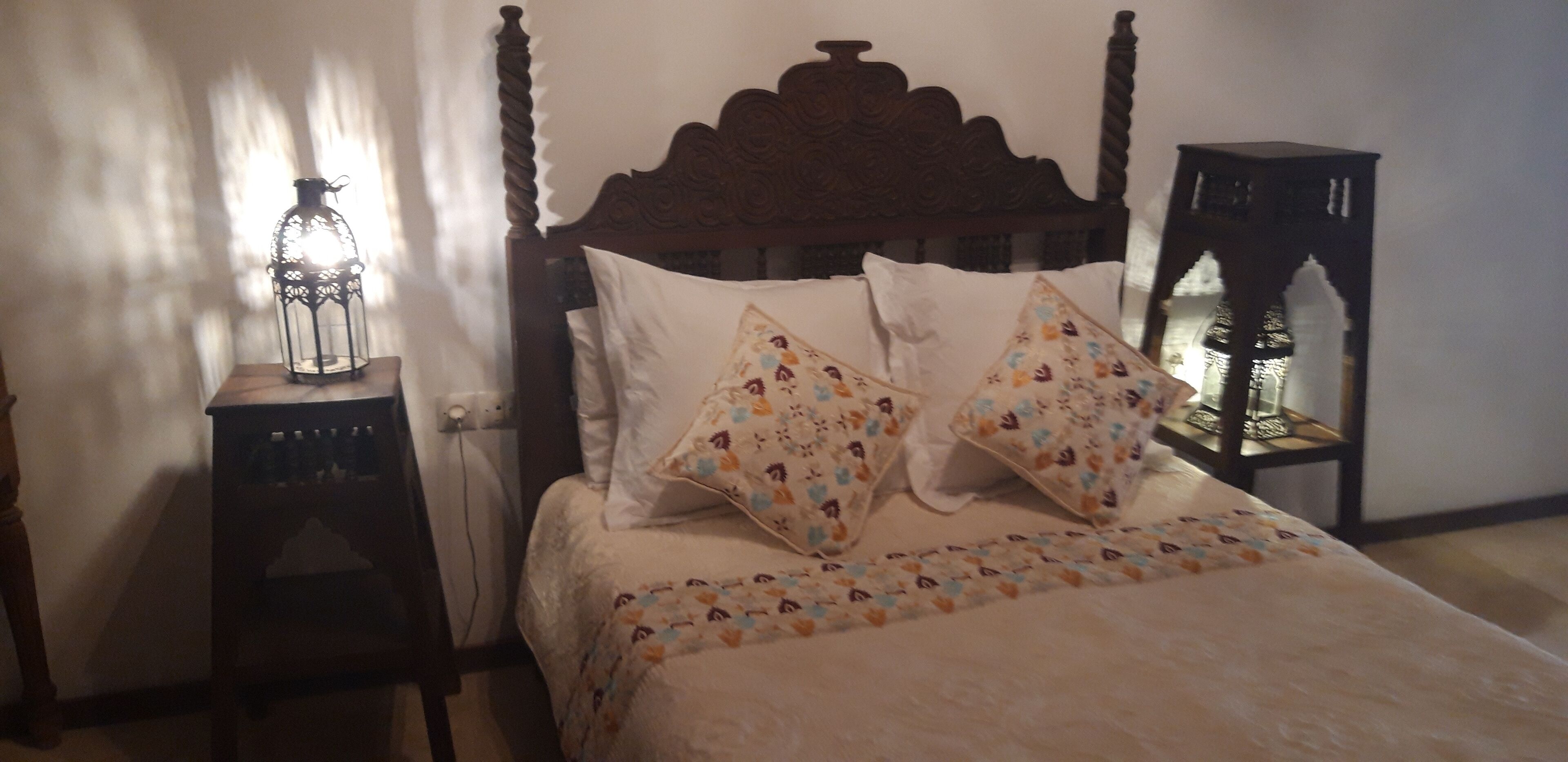Une chambre à coucher avec une tête de lit en bois, deux coussins à motifs, une table de chevet avec une lanterne et une petite table de nuit avec une lampe.