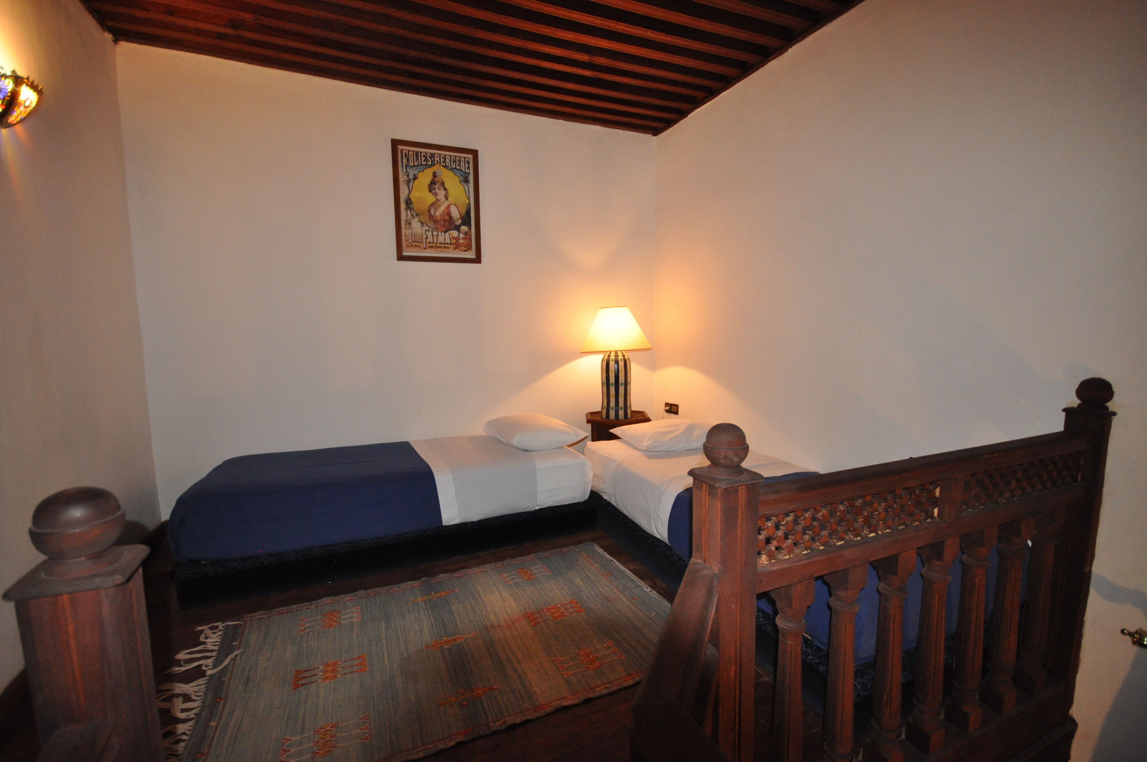 A room with two single beds, a wooden headboard, a nightstand, a lamp, a poster on the wall, and a patterned rug.