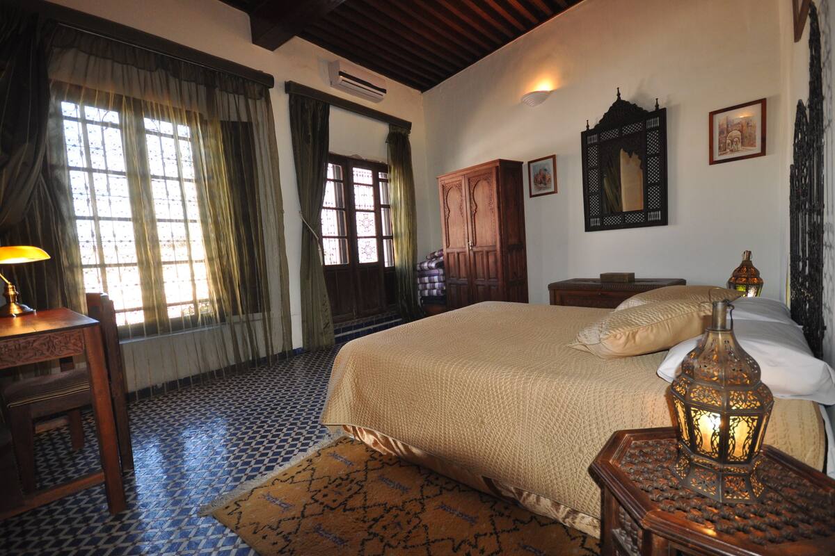 Quarto com uma cama grande, uma mesa de cabeceira de madeira, uma escrivaninha com abajur e um chão estampado.