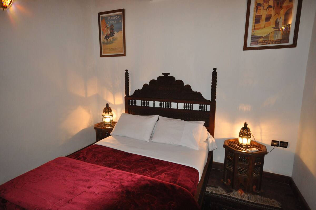 Uma cama com cabeceira de madeira, duas luminárias laterais e um cobreleito vermelho.