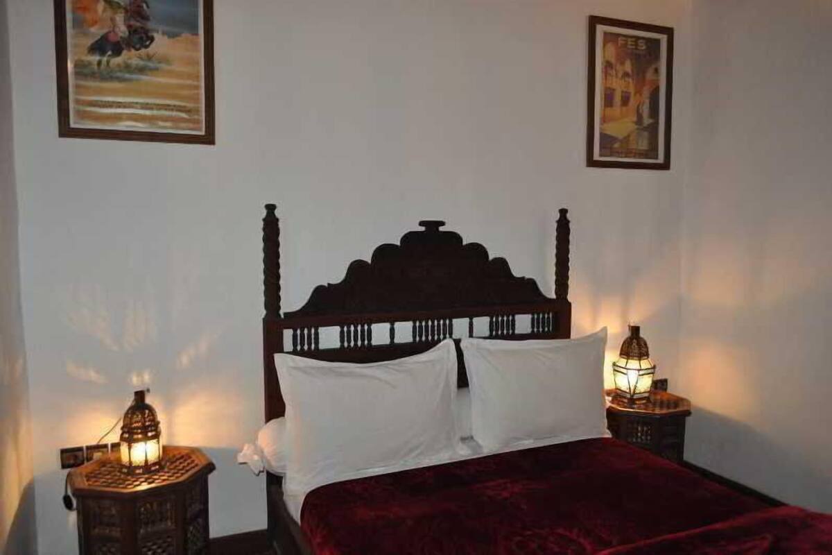 Uma cama com cabeceira de madeira escura, duas almofadas brancas e um cobertor vermelho. Há duas mesas de cabeceira fixadas na parede com luminárias em estilo de lanterna tradicional.