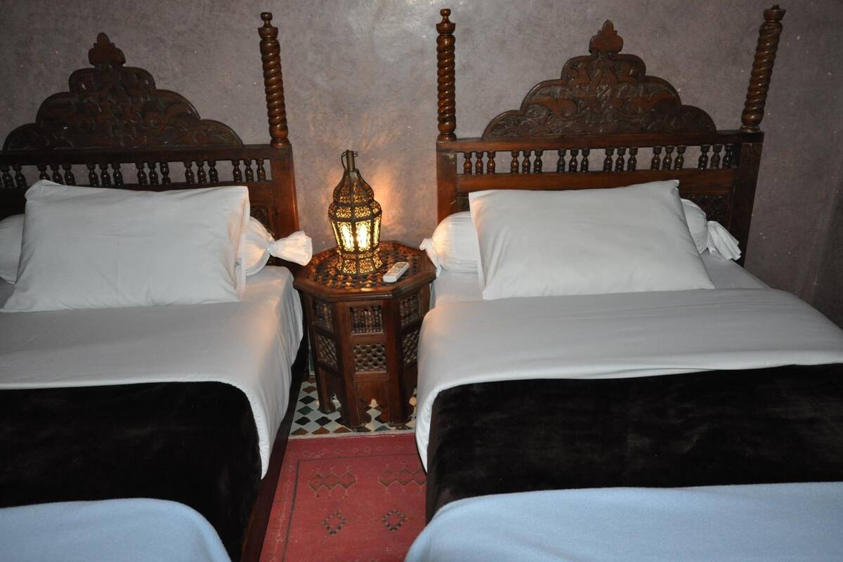 Quarto com duas camas, cada uma com cabeceira de madeira e roupa de cama branca. Pequeno criado-mudo de madeira com um candeeiro entre as camas.
