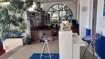 A modern interior with a blue carpet, two bar stools, a wicker sofa, and a large window with plants.