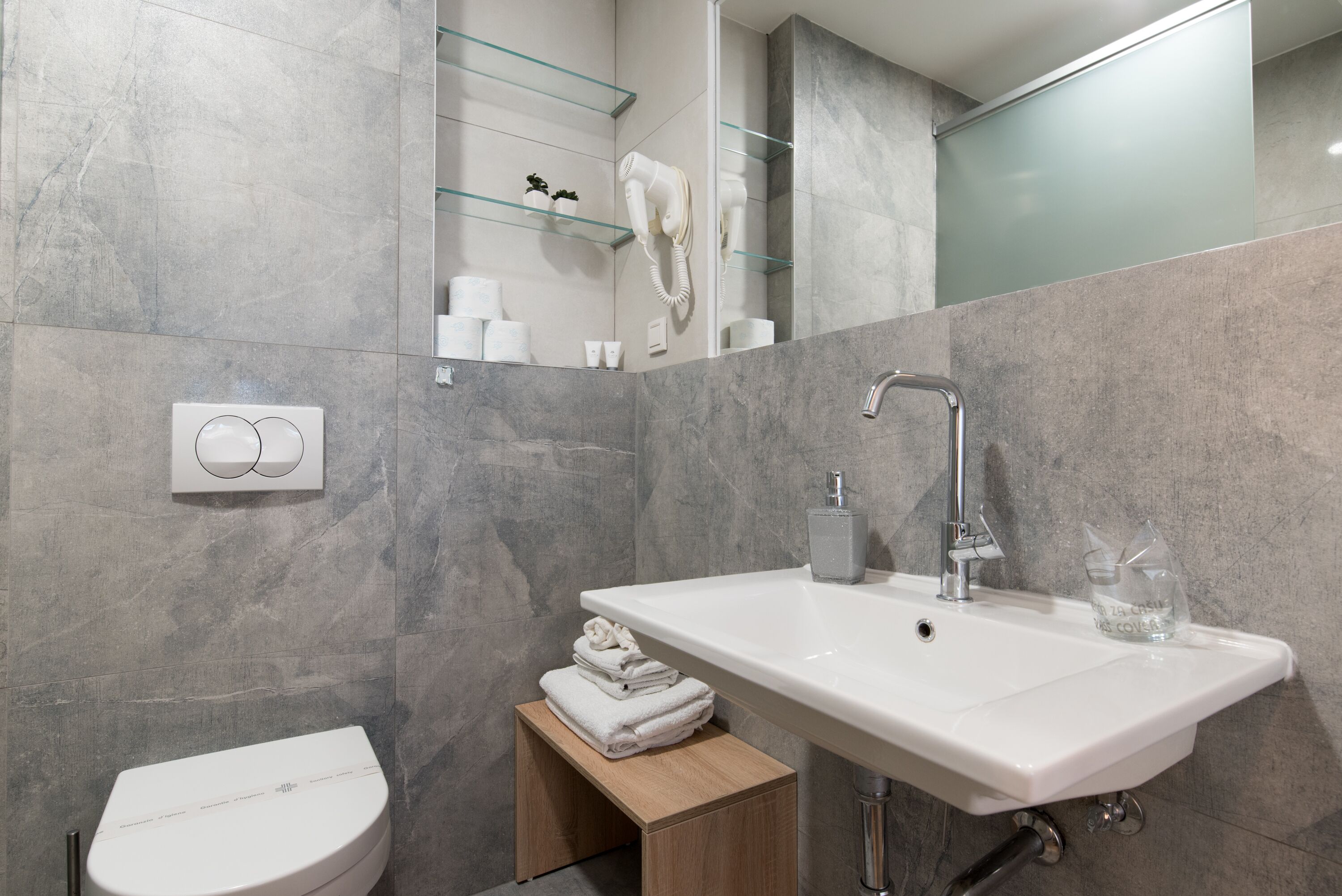 A modern bathroom with a white sink, a toilet, and a glass shelf with toiletries.