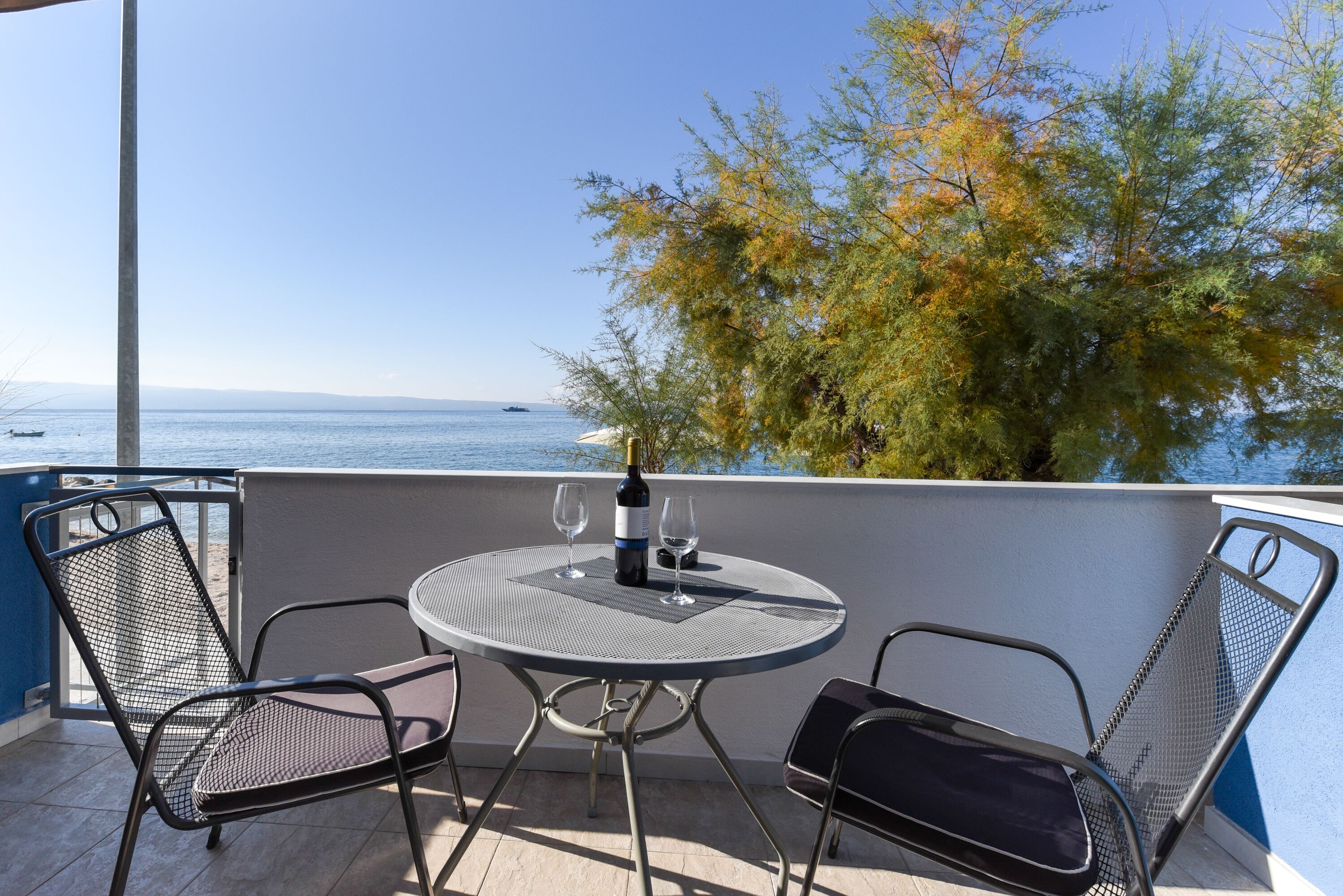 A balcony with a round table and two chairs, overlooking a body of water and a tree.