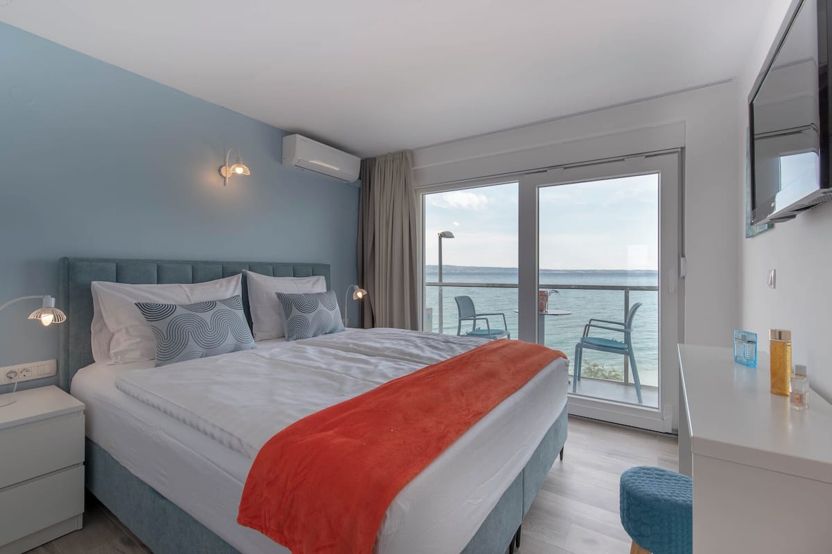 A hotel room with a bed, a nightstand, a desk, a chair, and a view of the sea through a large window.