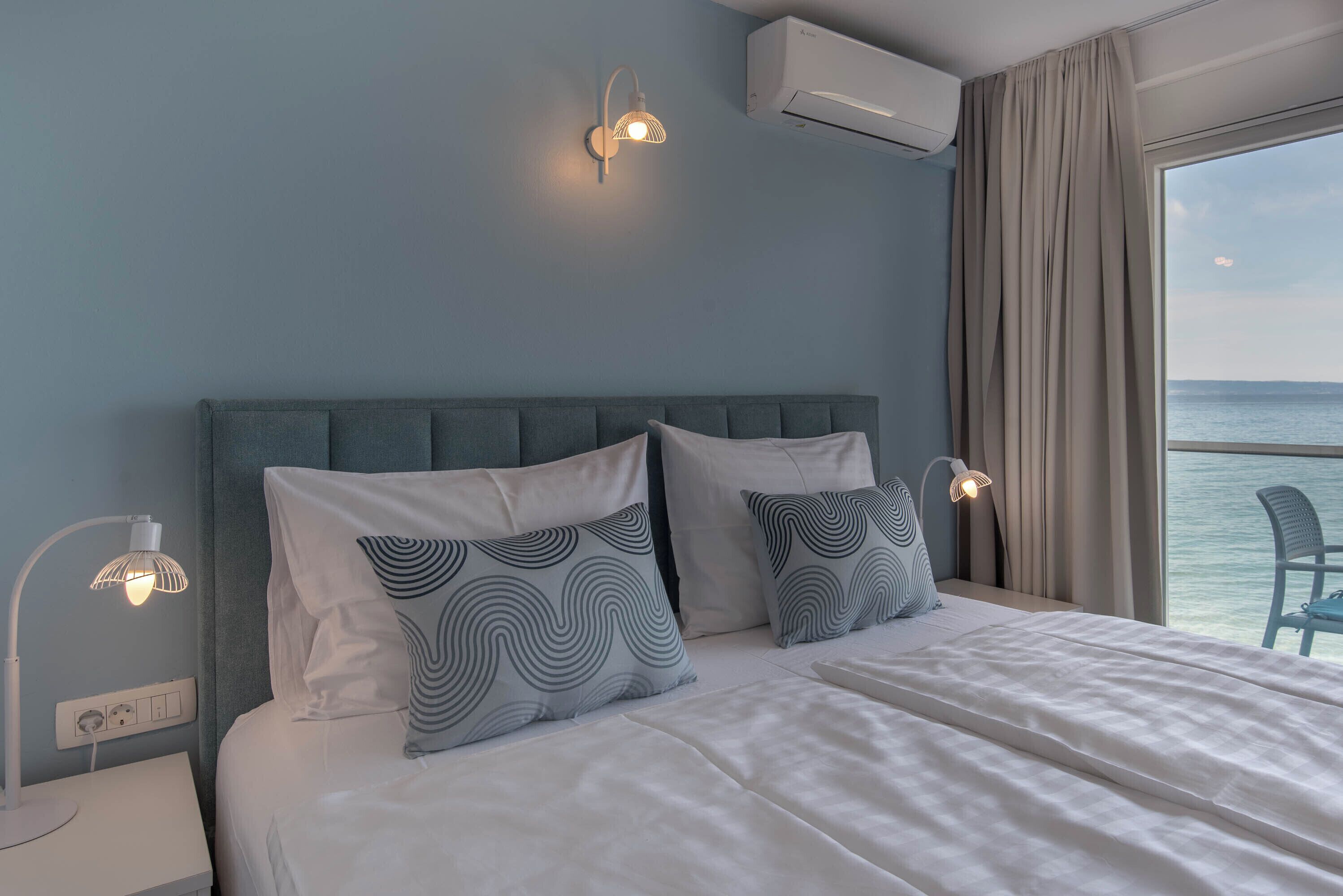 A neatly made bed with white linens and patterned pillows, a bedside table with a lamp, and a view of the sea through a large window.