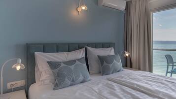 A neatly made bed with white linens and patterned pillows, a bedside table with a lamp, and a view of the sea through a large window.