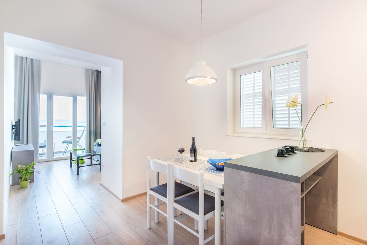A modern kitchen with a dining area, a large window with blinds, a hanging light fixture, and a potted plant.