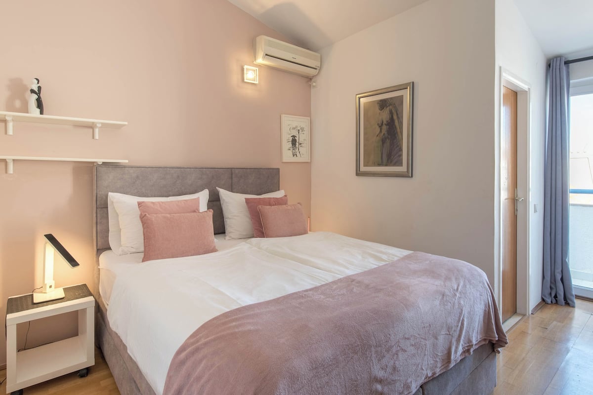A bedroom with a bed, bedside tables, a wall-mounted air conditioner, a framed picture, and a window with curtains.