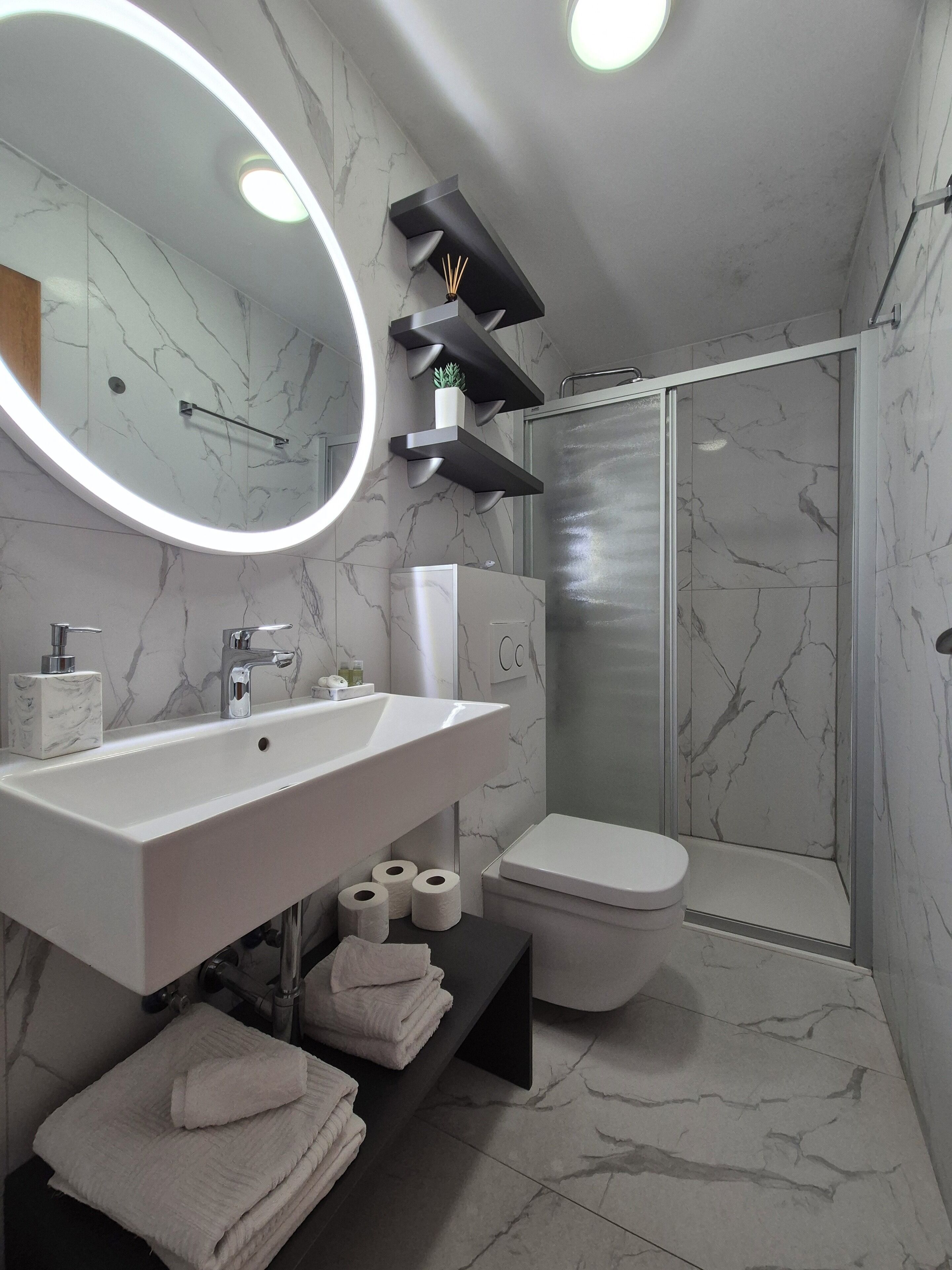 A modern bathroom with a white sink, mirror, and toilet, marble walls, and a glass shower enclosure.