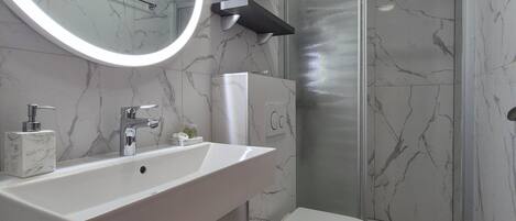 Une salle de bain moderne avec un lavabo, un miroir et des toilettes blancs, des murs en marbre et une cabine de douche en verre.