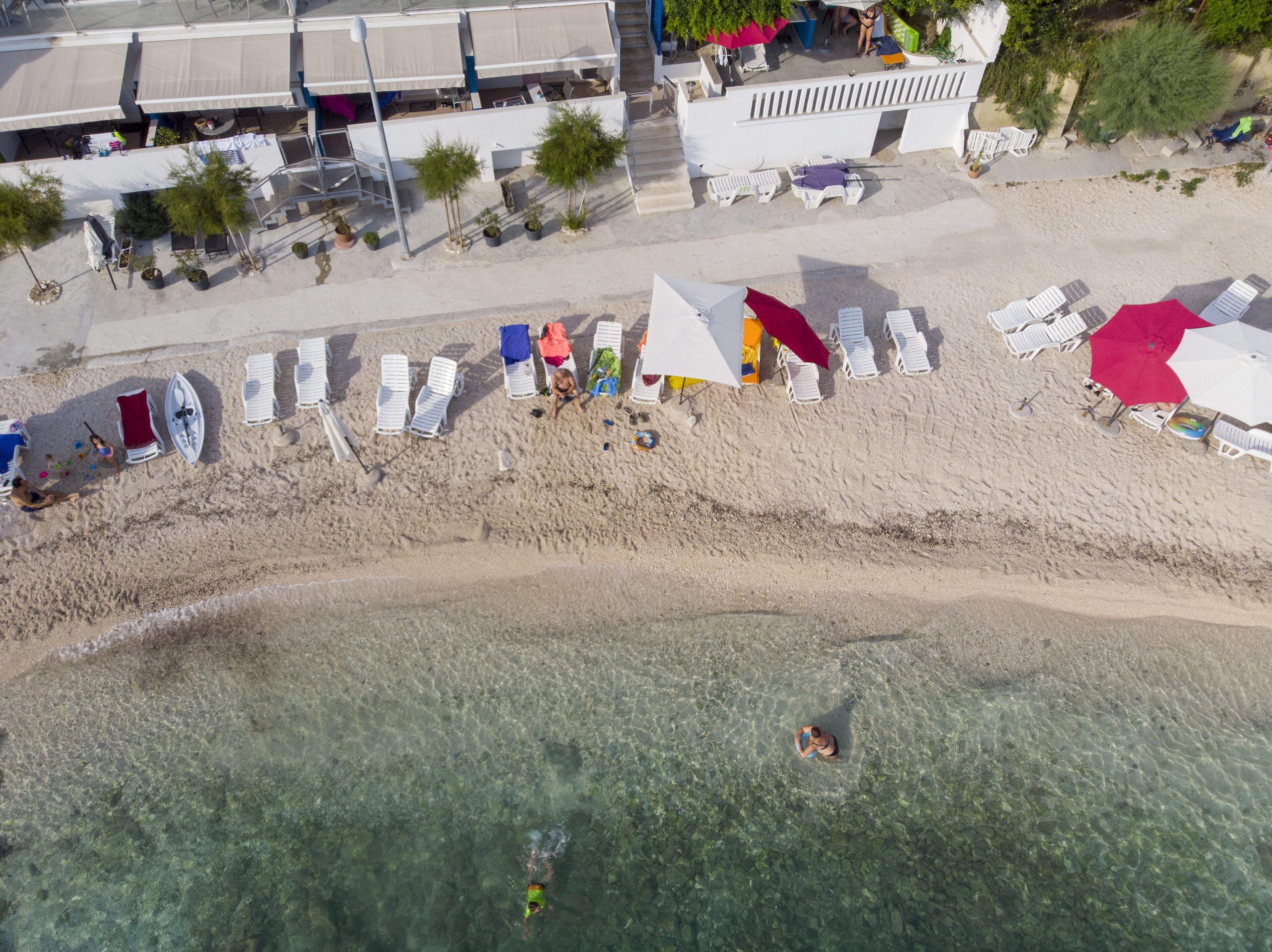 An aerial view of a beach with clear shallow waters, rows of sun loungers, and umbrellas.