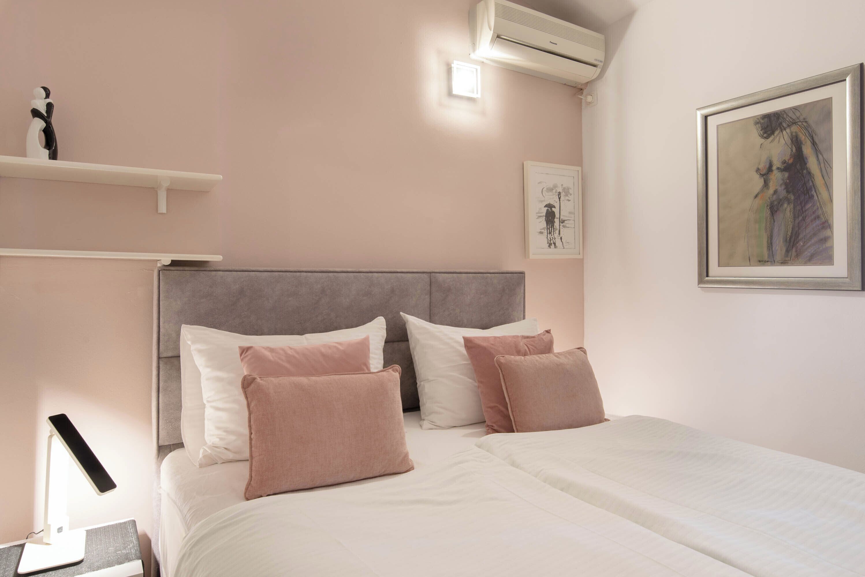 A neatly made bed with a grey headboard, white and pink pillows, and a bedside table with a lamp and a phone.