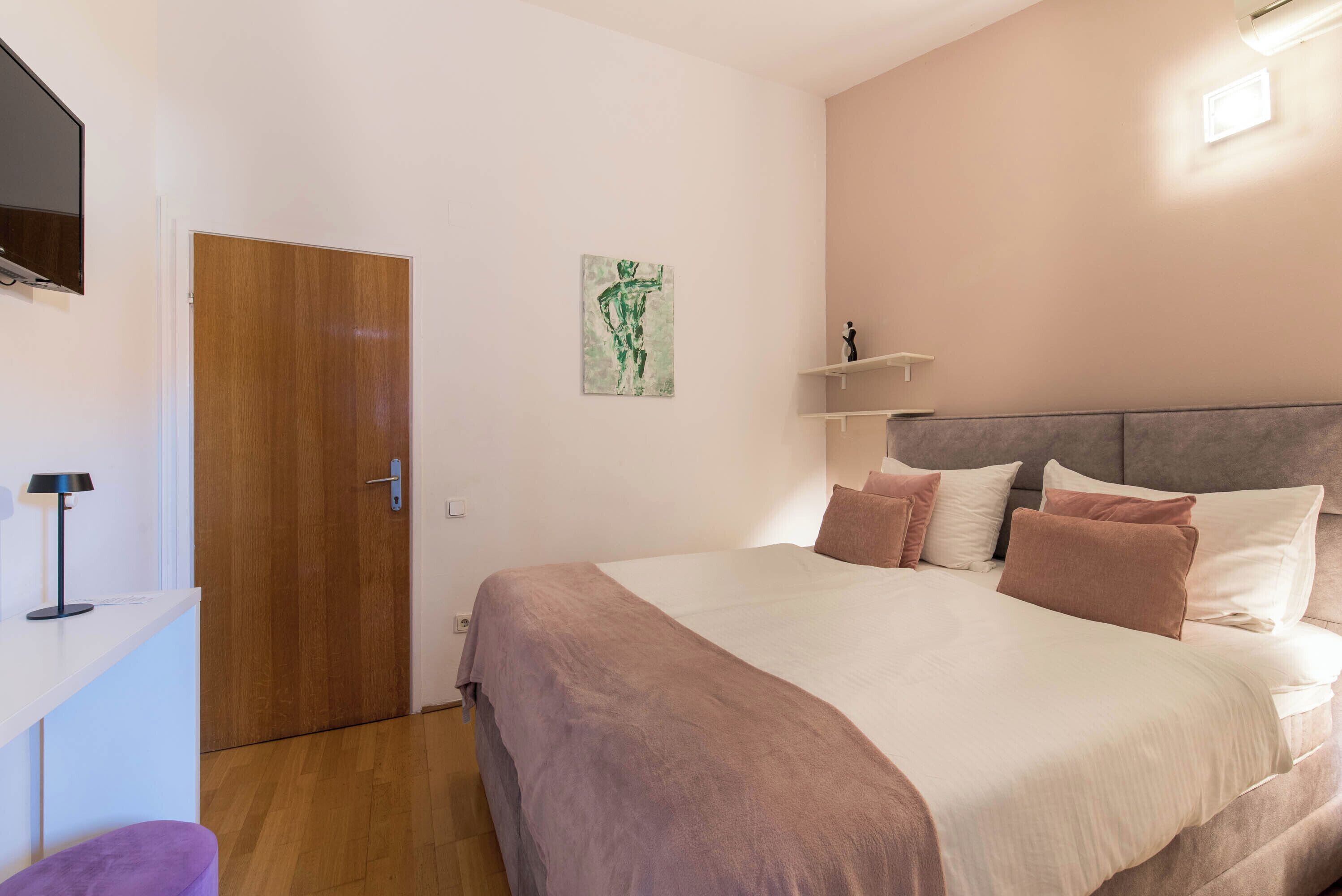 Ein Schlafzimmer mit einem Bett, einer Holztür, einem an der Wand befestigten Fernseher und einem Gemälde.