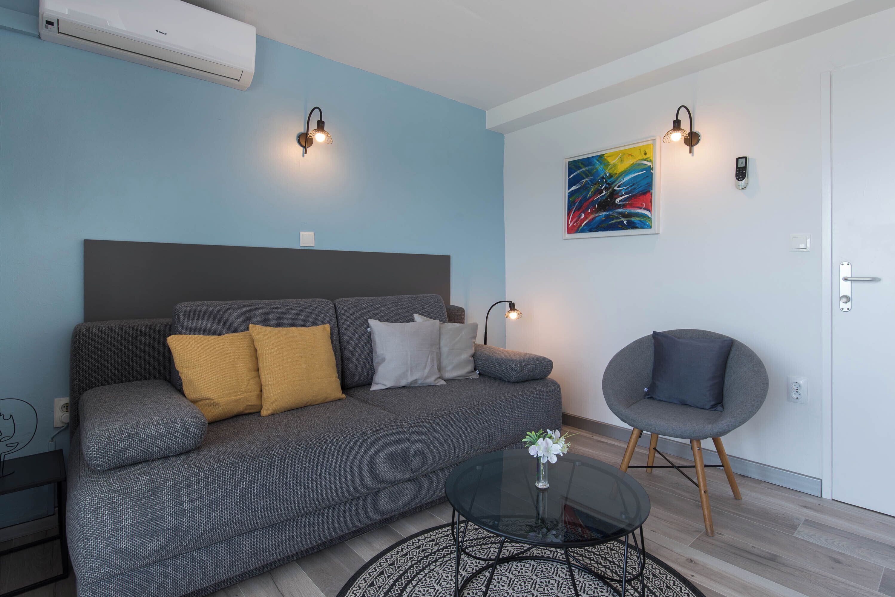 Uma sala de estar moderna com um sofá cinzento, uma mesa de centro redonda e uma poltrona cinzenta. Há um quadro na parede e um aparelho de ar condicionado fixado na parede.